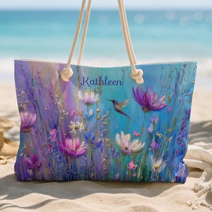 May include: A large tote bag with a floral design featuring purple, pink, and white flowers, and a hummingbird. The bag has cream-colored rope handles and the name "Kathleen" is printed on the front. The bag is on a sandy beach.