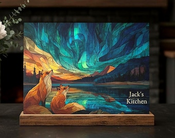 Peut inclure: Un panneau rectangulaire en bois présentant une illustration style vitrail de deux renards observant une aurore boréale bleue et verte au-dessus d'un lac. Le texte "Jack's Kitchen" est imprimé en blanc sur le côté inférieur droit du panneau.