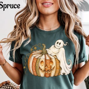 May include: A dark green short-sleeve t-shirt featuring a graphic of a pumpkin made of stacked books with a smiling ghost draped over the top. The text Blue Spruce is printed in the upper left corner of the image in a black serif font.