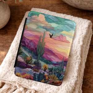 May include: A rectangular e-reader case featuring a colorful desert landscape illustration. The design displays a saguaro cactus, smaller desert plants, and a bird silhouette against a vibrant sky of layered pink, purple, yellow, and teal clouds.