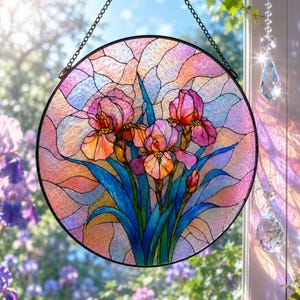 May include: A circular stained glass suncatcher featuring three blooming iris flowers in shades of pink, orange, and yellow, with deep blue leaves. The piece is framed in black metal and hangs from a black chain against a bright window background.