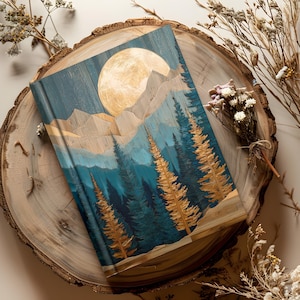 May include: A hardback journal with a landscape design featuring a full moon, mountains, and evergreen trees. The cover has a blue and gold color scheme, with a wood-grain texture. The journal is resting on a wooden slice.