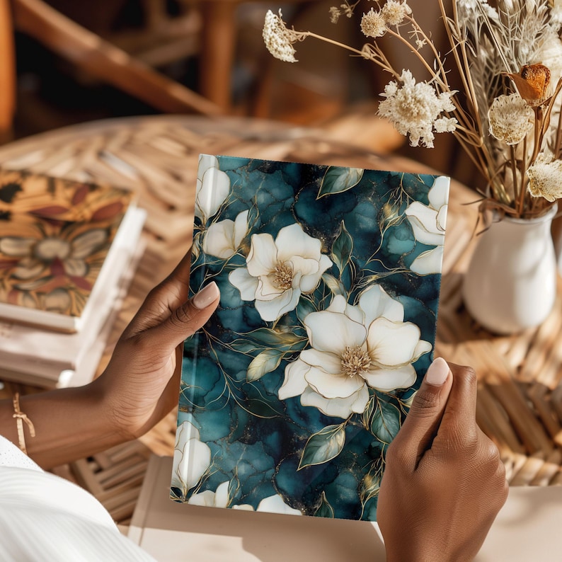 May include: A floral journal with a teal and gold marble design. The cover features large white flowers with gold outlines and green leaves. The journal is held open, showcasing the detailed floral pattern.