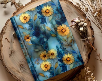 May include: A hardcover journal with a floral design featuring yellow daisies and blue foliage on a light blue background. The journal is resting on a wooden slice, with dried flowers and branches around it.