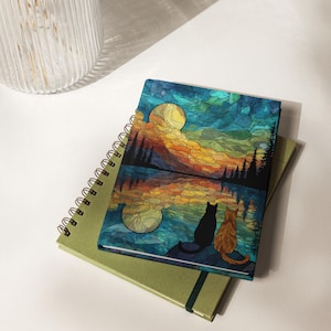May include: A rectangular notebook featuring a stained-glass style illustration of a black cat and an orange cat sitting by a lake at night. The scene depicts a large yellow moon, dark silhouetted pine trees, and a reflection on the water surface.