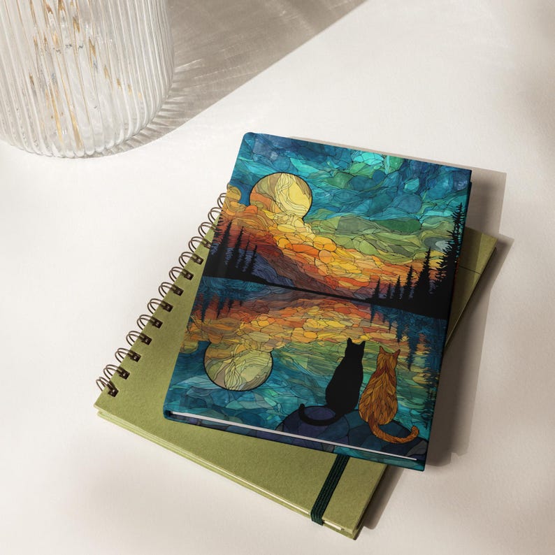 May include: A rectangular notebook featuring a stained-glass style illustration of a black cat and an orange cat sitting by a lake at night. The scene depicts a large yellow moon, dark silhouetted pine trees, and a reflection on the water surface.