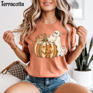 May include: A terracotta-colored short-sleeve t-shirt featuring a graphic print of a carved pumpkin made of stacked books with a white ghost draped over the top. The pumpkin has a smiling face and intricate decorative patterns on its segments.