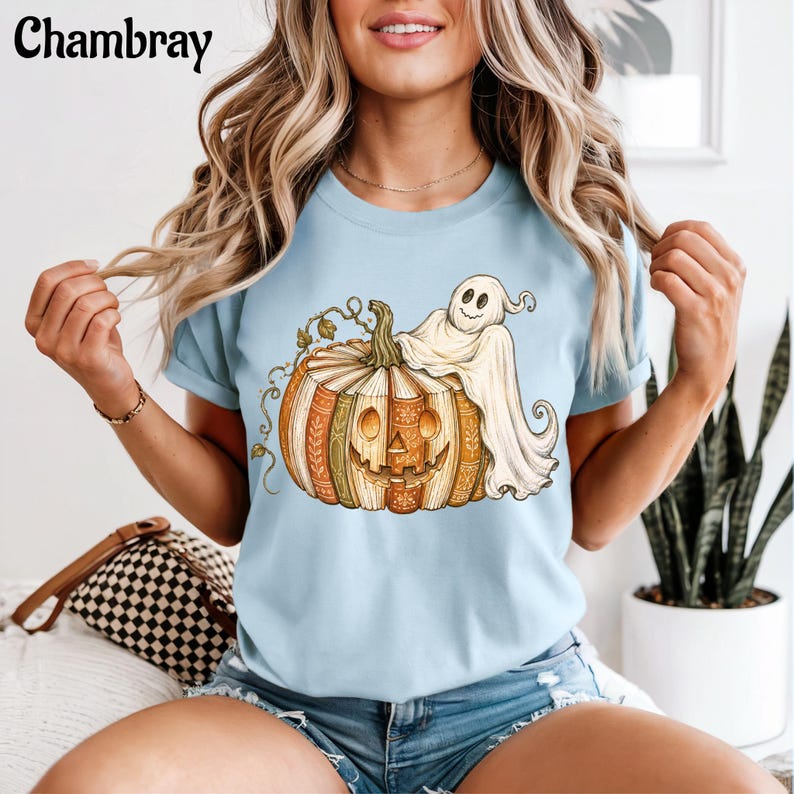 May include: A light blue t-shirt featuring a graphic of a jack-o'-lantern made of stacked books with a ghost draped over the top. The pumpkin has a carved face and intricate spine details. The word Chambray is printed in the top left corner.
