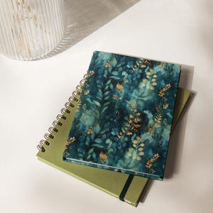 May include: A spiral-bound notebook with a dark teal and blue watercolor background featuring gold botanical leaf illustrations, resting on top of a solid olive green notebook with a black elastic closure band and a black wire spiral binding.