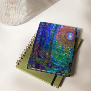 May include: A rectangular notebook featuring a vibrant, multi-colored cover with a mosaic-like pattern of deep blues, greens, and a central sun-like circular design, resting on top of a larger, plain olive-green spiral-bound notebook.
