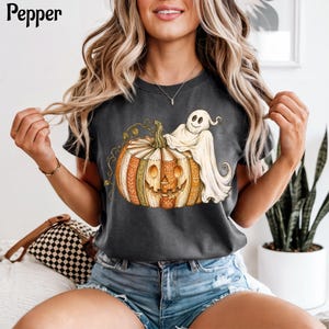 May include: A charcoal gray t-shirt featuring a graphic of a jack-o'-lantern made of stacked books with a white ghost draped over the top. The word Pepper is printed in black in the upper left corner. The shirt has short sleeves and a crew neck.