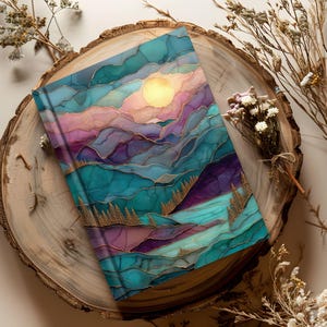 May include: A hardback journal with a colorful abstract landscape design. The cover features a mountain scene with teal, purple, and pink hues, a golden sun, and gold accents. The journal is resting on a wooden slice.