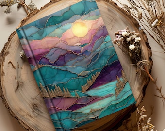 May include: A hardback journal with a colorful abstract landscape design. The cover features a mountain scene with teal, purple, and pink hues, a golden sun, and gold accents. The journal is resting on a wooden slice.