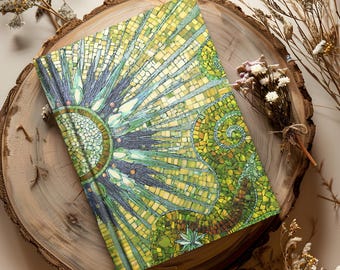 May include: A decorative journal with a mosaic-style design featuring shades of green, yellow, and blue. The cover has a sunburst pattern and a spiral design. The journal is resting on a wooden surface.