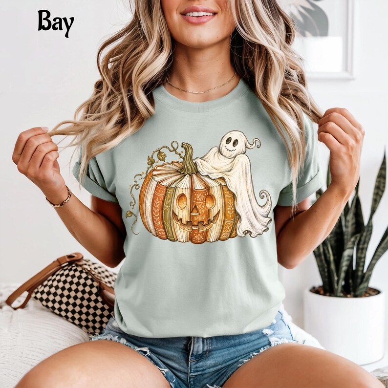 May include: A light green t-shirt featuring a graphic of a jack-o'-lantern made of stacked books with a white ghost draped over the top. The text "Bay" is printed in black in the upper left corner. The shirt has short sleeves and a crew neck.