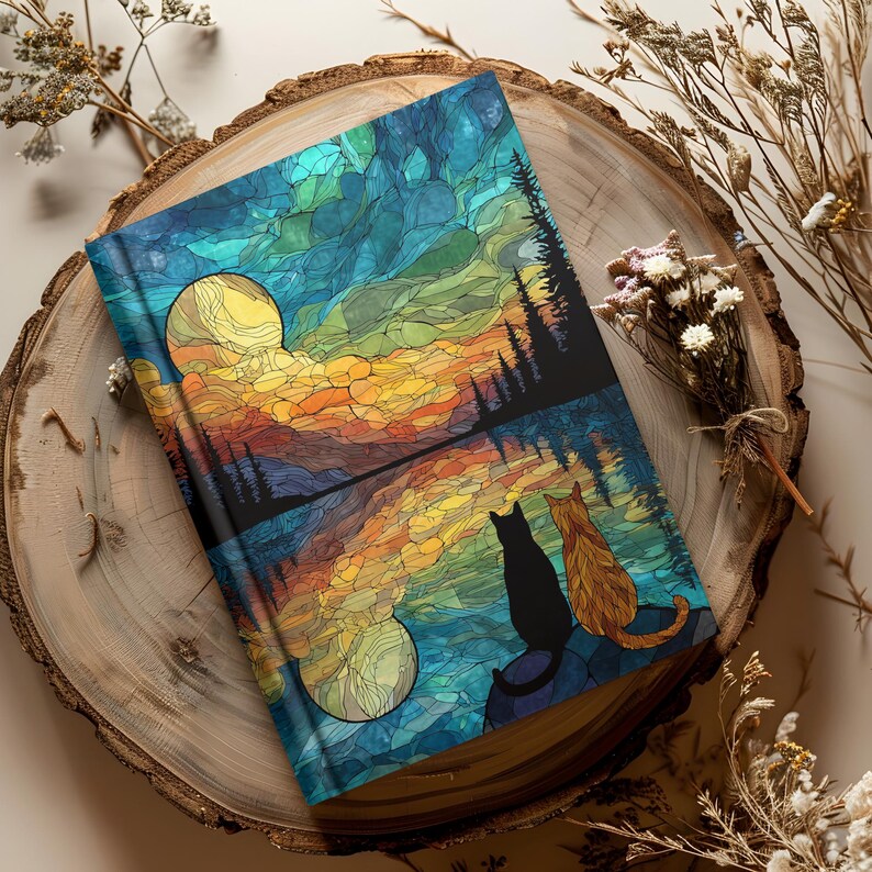 May include: A hardcover journal featuring a stained-glass style illustration of a black cat and an orange tabby cat sitting together, looking out at a lake reflecting a vibrant sunset with a large yellow moon against a deep blue sky.