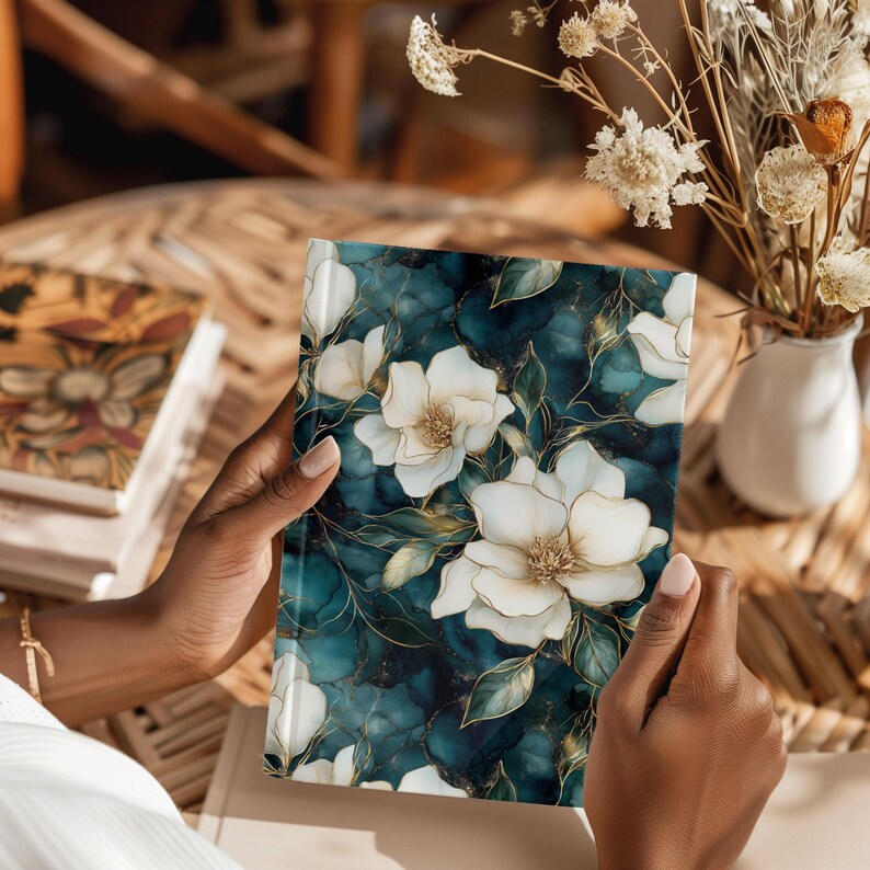 May include: A floral journal with a teal and gold marble design. The cover features large white flowers with gold outlines and green leaves. The journal is held open, showcasing the detailed floral pattern.