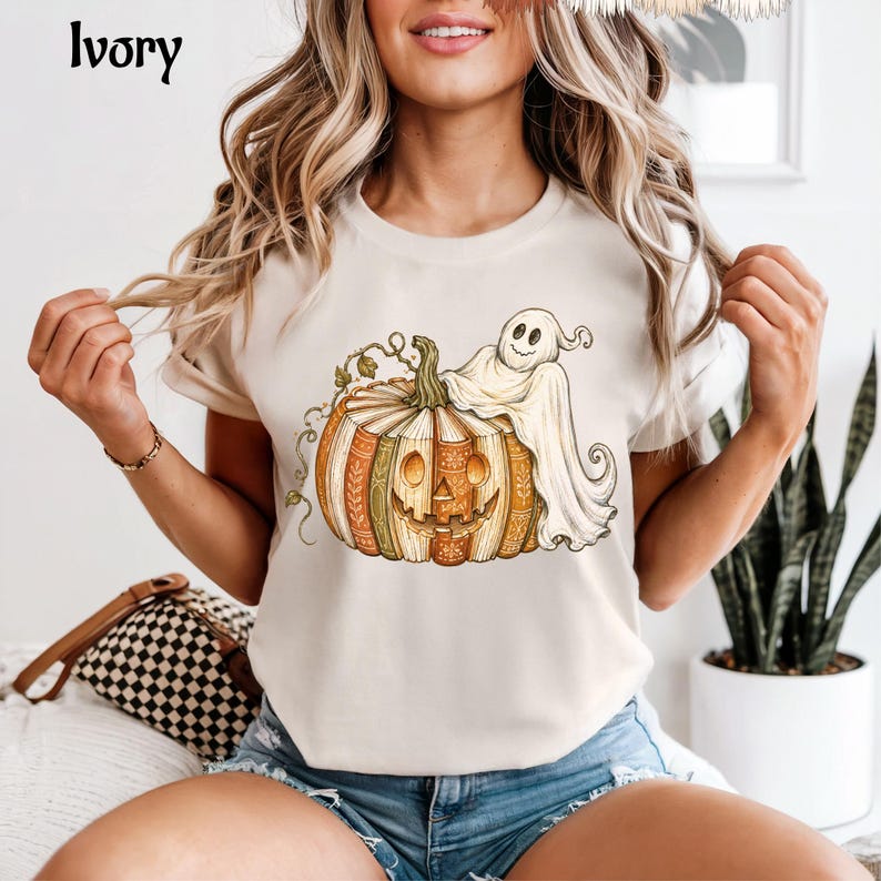 May include: An ivory short-sleeve t-shirt featuring a graphic print of a carved pumpkin made of stacked books with a ghost draped over the top. The pumpkin has a smiling face and the books have orange, green, and brown spines with gold details.