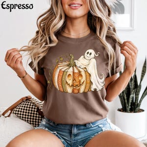 May include: A brown short-sleeve t-shirt featuring a graphic print of a jack-o'-lantern pumpkin made of stacked books with a white ghost draped over the top. The word Espresso is printed in black in the upper left corner of the image.