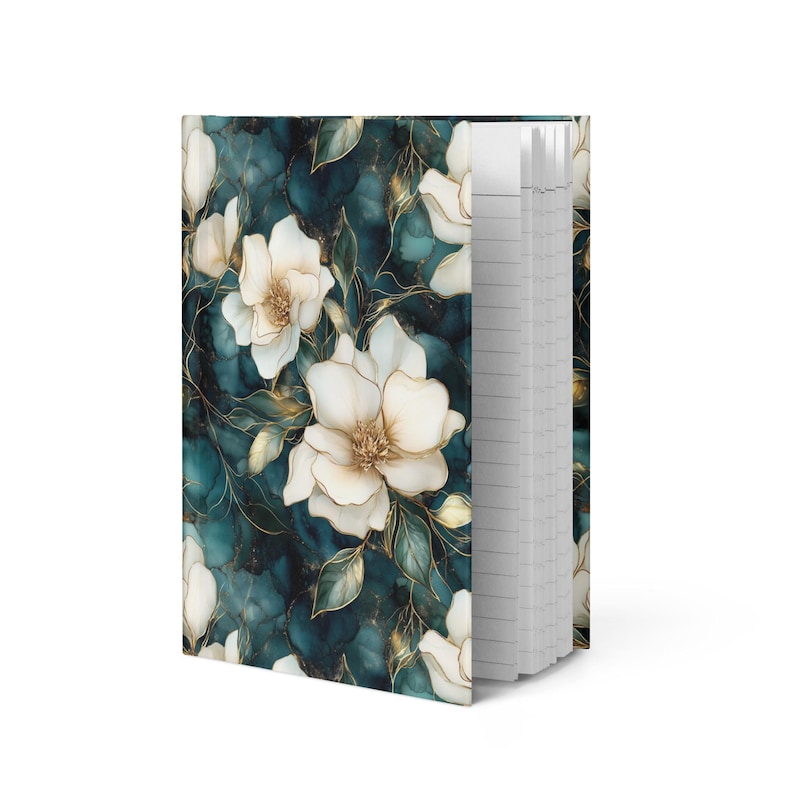 May include: A hardcover journal with a floral design. The cover features white flowers with gold accents against a teal and dark blue background. The journal's pages are lined and visible on the right side.