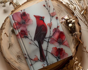 May include: A decorative journal featuring a vibrant red cardinal perched on a flowering branch. The cover art showcases watercolor-style red flowers and a bird against a soft, light background. The journal is displayed on a wooden slice.