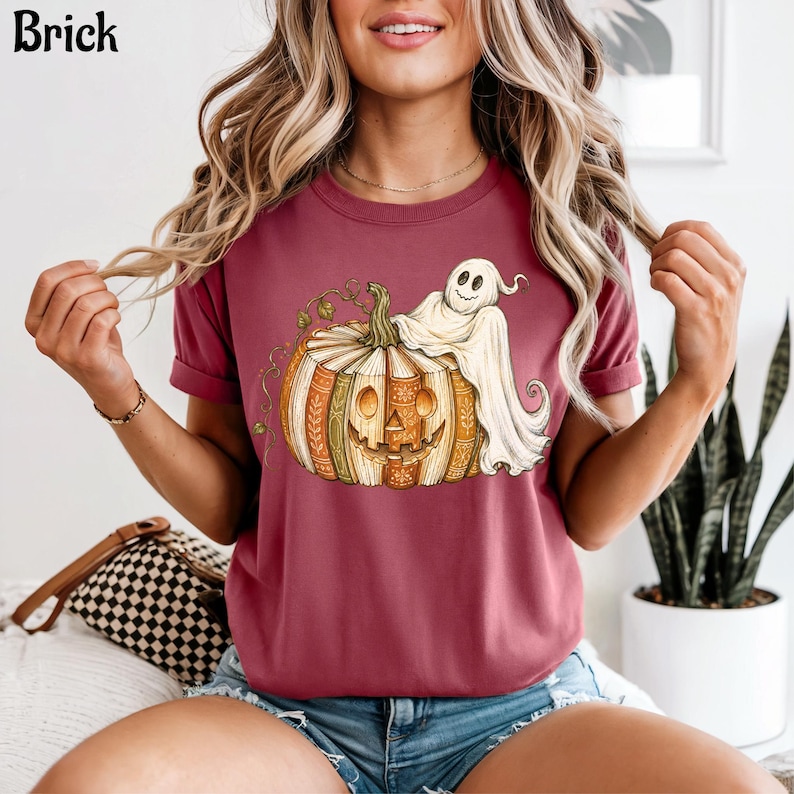 May include: A brick-colored short-sleeve t-shirt featuring a graphic print of a carved pumpkin with a book-like texture and a white ghost draped over the top. The pumpkin has a smiling face and green vines, while the ghost has a simple face.
