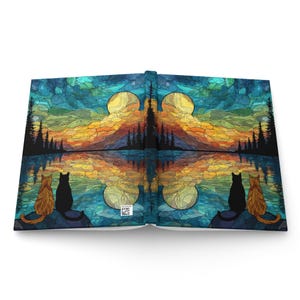 May include: A hardcover journal featuring a stained-glass style landscape with a sunset, silhouetted pine trees, and a reflective lake. Two cats, one orange and one black, sit on each side of the cover, looking toward the horizon.