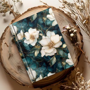 May include: A floral journal with a teal and gold marble design. The cover features large white flowers with gold accents and green leaves. The journal is displayed on a wooden slice with dried flowers.