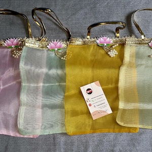 May include: Four rectangular sheer fabric bags in light pink, pale green, mustard yellow, and pale yellow. Each bag features gold metallic handles, a gold sequin trim, and a pink lotus flower embellishment with a cluster of small white faux pearls.