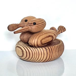 May include: A carved wooden sculpture featuring two duck-like birds resting inside a circular wooden bowl. The wood displays prominent, swirling grain patterns in light and dark brown tones, with small black beads serving as the eyes for each bird.