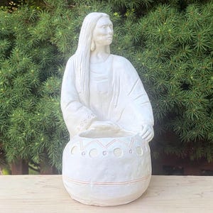 May include: A white ceramic sculpture of a person with long hair wearing a robe, positioned behind a rounded vessel. The vessel features a decorative pattern of horizontal lines, small circles, and zig-zag shapes etched into the surface.