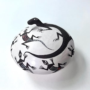 May include: A round, white ceramic vessel featuring a hand-painted black lizard sculpture attached to the top. The surface is decorated with multiple black and white lizard illustrations, each containing intricate geometric patterns and small red accents.