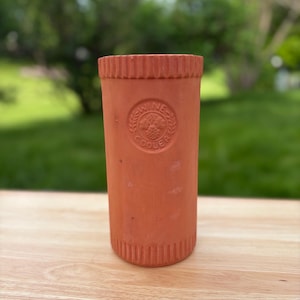 May include: A terracotta wine cooler with a ridged top and a circular emblem that reads "Wine Cooler". The cylindrical container is a warm reddish-brown color and sits on a wooden surface. The background is blurred with green foliage.