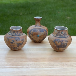 May include: Three tan ceramic vessels with narrow necks and wide bodies. Each piece features dark brown etched animal figures, geometric patterns, and a small, circular, bright blue stone inlay centered on the front of each pot.