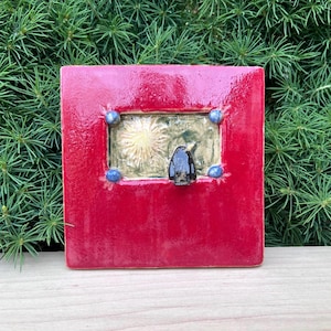 May include: A square, red ceramic tile with a recessed, gold-colored center featuring a bird figure. The tile is adorned with four blue, spherical accents. The background is a lush green, creating a striking contrast.
