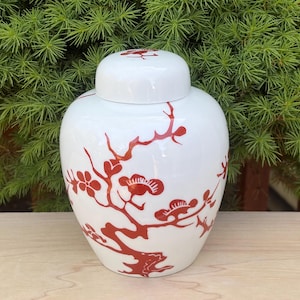 May include: A white ceramic ginger jar with a matching lid. The jar is decorated with a red floral and branch design. The jar is sitting on a light brown wooden surface with green foliage in the background.