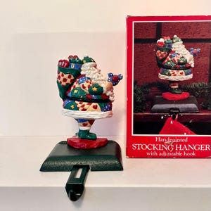 May include: A handpainted Santa Claus stocking hanger with an adjustable hook. The Santa figure wears a colorful patchwork coat with stars and hearts, carries a green sack of gifts, and holds a blue bird. The base is dark green metal.