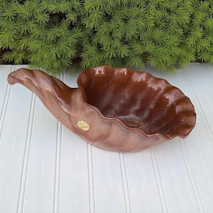 May include: A brown ceramic cornucopia-shaped bowl with a ruffled rim. The bowl has a glossy finish and a small, round sticker on the side. The background features green foliage and a white-painted wooden surface.
