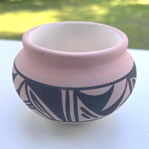 May include: A small, round, ceramic pot with a light pink exterior and a white interior. The pot features a black and cream geometric pattern around the base. The pot is likely handmade and suitable for decorative purposes.