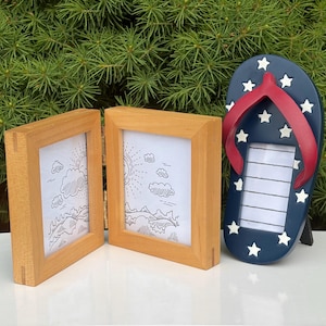 May include: Two light-colored wooden rectangular picture frames hinged together, standing next to a dark blue flip-flop shaped picture frame with a red strap and white star accents. The frames contain white inserts with line art drawings.