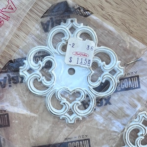 May include: White decorative hardware with an ornate, symmetrical design. The hardware has a central hole and is packaged in a clear plastic bag. The tag reads "True Value".