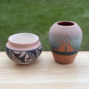 May include: A small, round, light pink bowl with black geometric patterns and a taller, rounded terracotta vase featuring a gradient of pink to grey with a dark horizontal band and a central orange geometric design depicting a stepped structure.