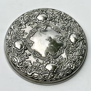 May include: A round silver-toned compact case featuring an intricate embossed floral and scrollwork pattern surrounding a smooth, reflective central area. The metallic surface displays detailed relief carvings of small flowers and leaves.