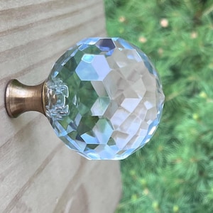 May include: A faceted crystal ball cabinet knob with a brass base. The clear, iridescent glass reflects light, creating a sparkling effect. The knob is mounted on a light-colored wooden surface, with a blurred green background.