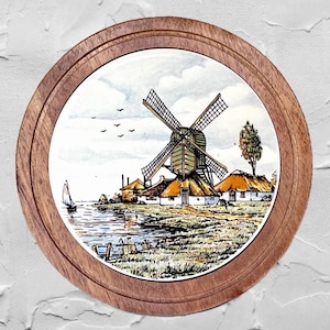 May include: Round wooden framed decorative plate featuring a watercolor painting of a Dutch windmill scene. The artwork depicts a windmill, houses, a sailboat, and birds against a cloudy sky. The frame is made of light brown wood.
