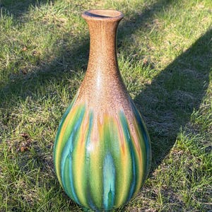 May include: A tall, decorative vase with a brown speckled top and a vibrant, colorful base. The base features vertical stripes of green, yellow, and blue. The vase is set on a bed of green grass.