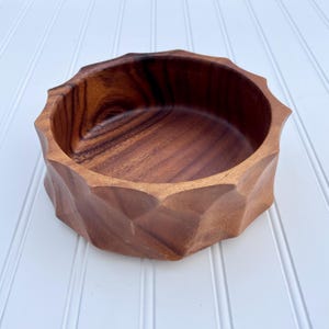 May include: A round wooden bowl featuring a dark brown grain pattern and a faceted exterior design with multiple angular, geometric edges around the rim and sides, creating a textured appearance with a smooth, polished interior surface.