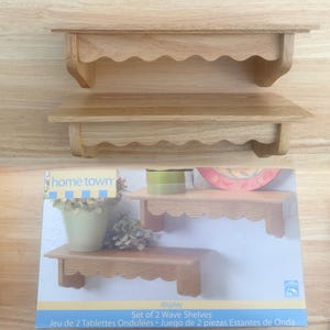 May include: A set of two light-colored wooden wall shelves featuring a scalloped, wave-like edge design on the front support brackets. The packaging box displays the product name home town and the text Set of 2 Wave Shelves.