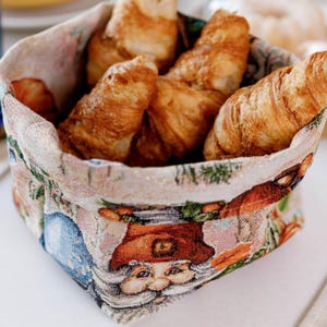 May include: A woven basket filled with golden-brown croissants. The basket has a festive design with a Santa Claus face and other holiday imagery. The basket is square-shaped and appears to be made of fabric.