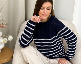 May include: A navy blue sweater with horizontal white stripes on the sleeves and lower torso. The sweater has a crew neck and is paired with cream-colored pants. The person is seated in a beige armchair.